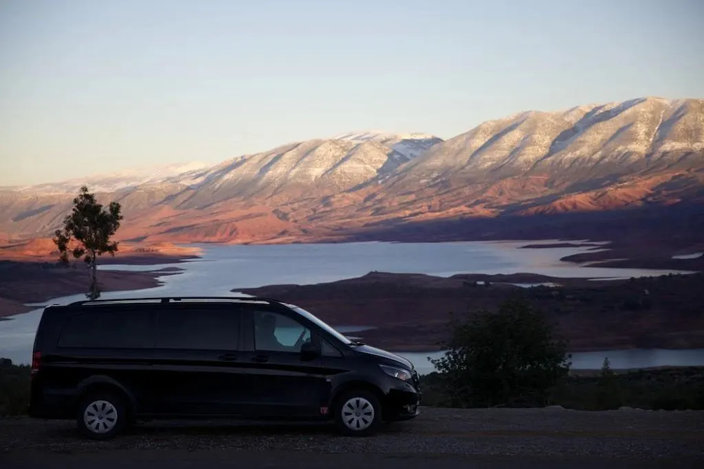 A black luxury passenger van parked on a scenic mountain overlook with a lake and snow-capped peaks