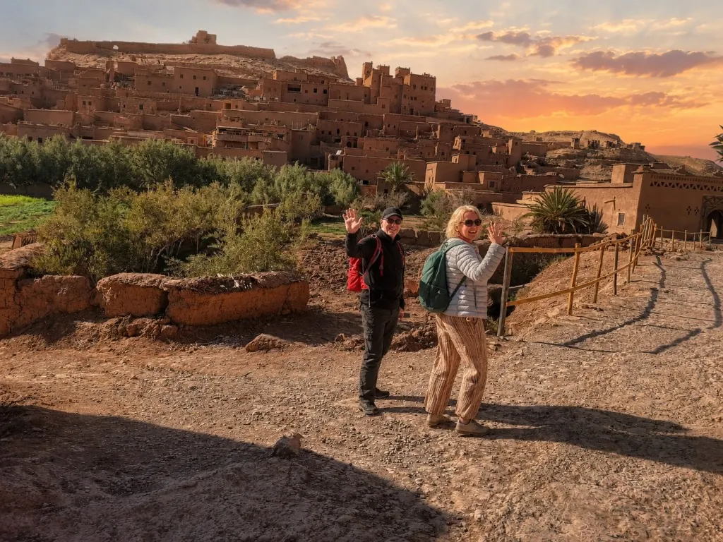 Two travellers waving in front of the UNESCO Kasbah of Ait Ben Haddou at sunset