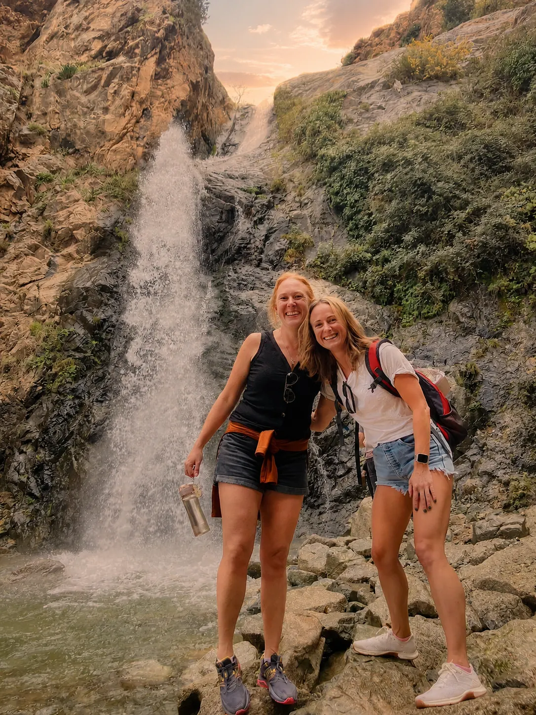 Two travellers smiling together at the base of a waterfall