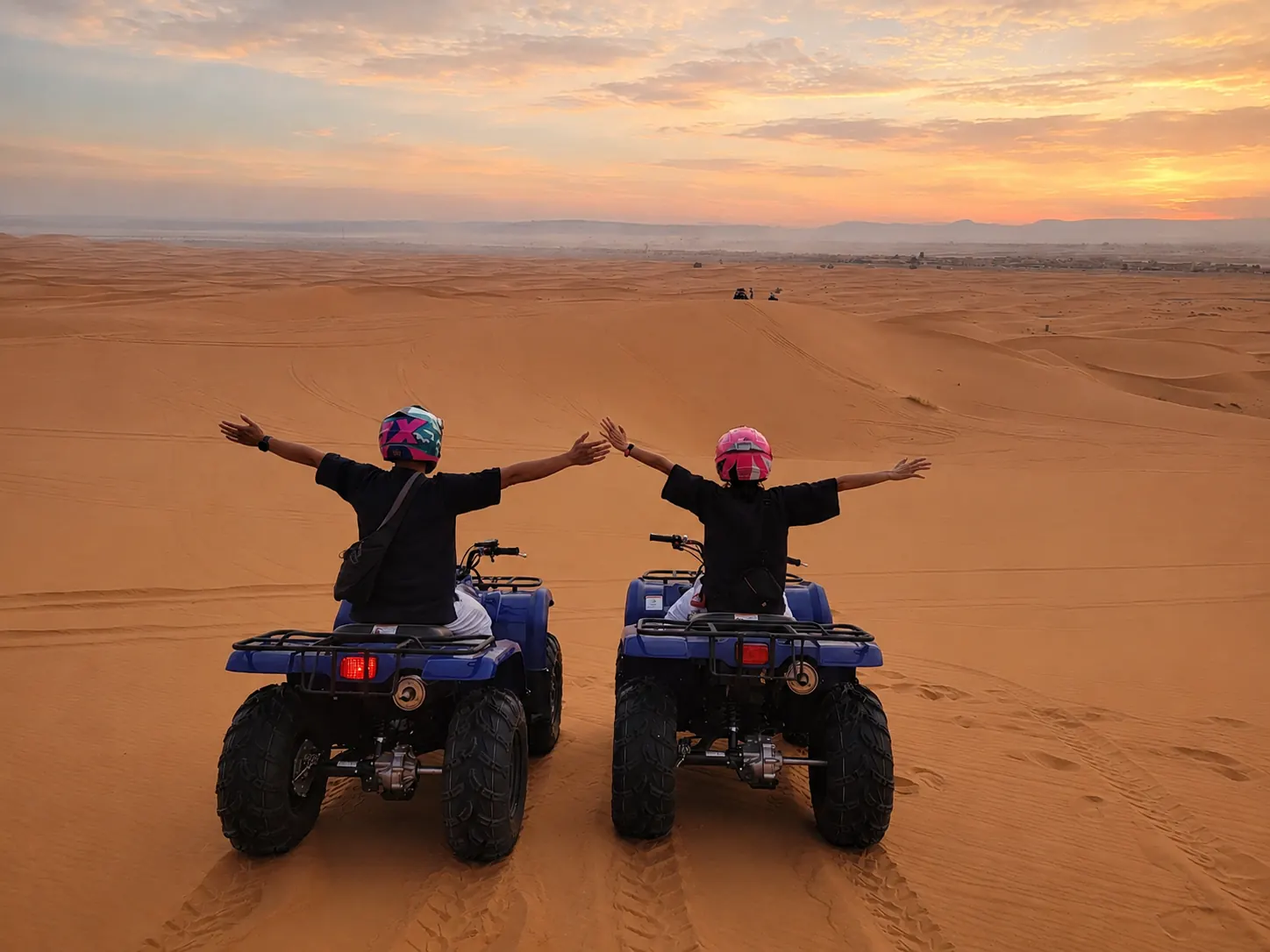Two quad bikers with arms outstretched against a sweeping Agafay Desert sunset